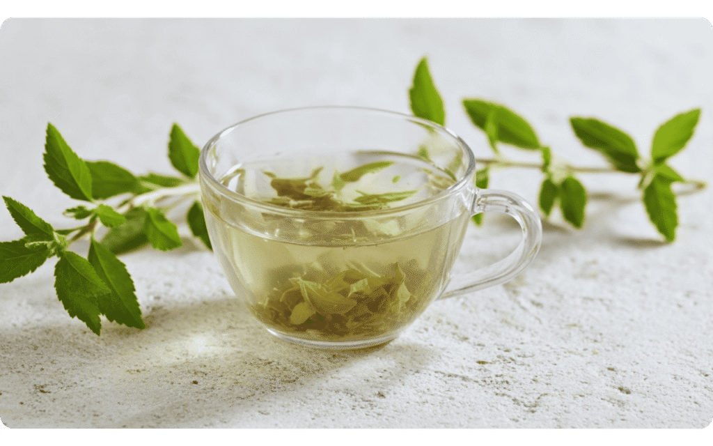 Xícara de vidro com chá verde e folhas frescas ao fundo sobre superfície clara.