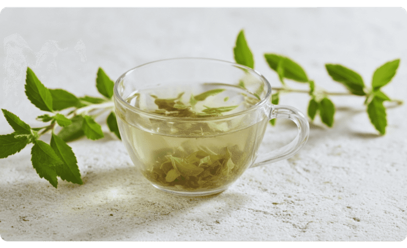 Xícara de vidro com chá verde e folhas frescas ao fundo sobre superfície clara.