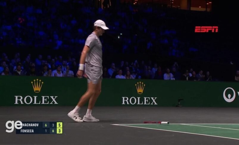 Quadra de tênis, jogador em ação e placar exibido ao fundo.