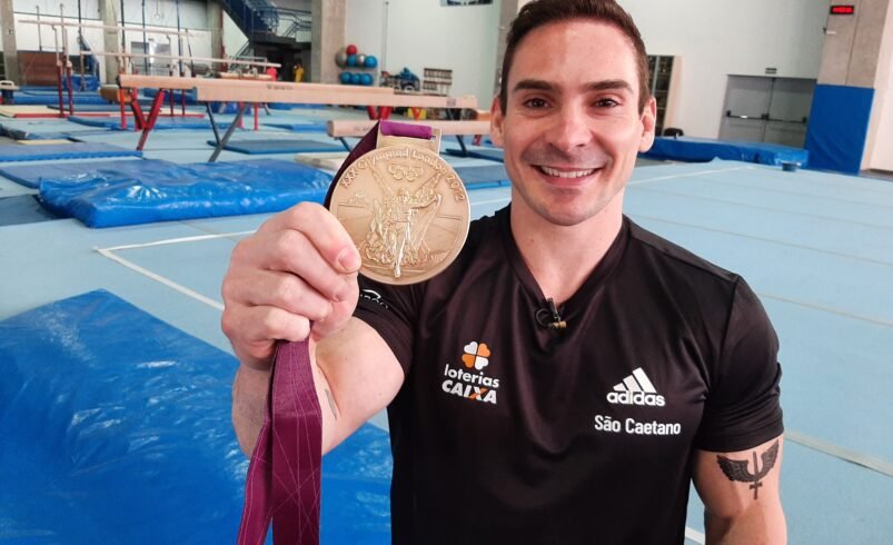 Pessoa segurando medalha dourada com equipamentos de ginástica ao fundo.