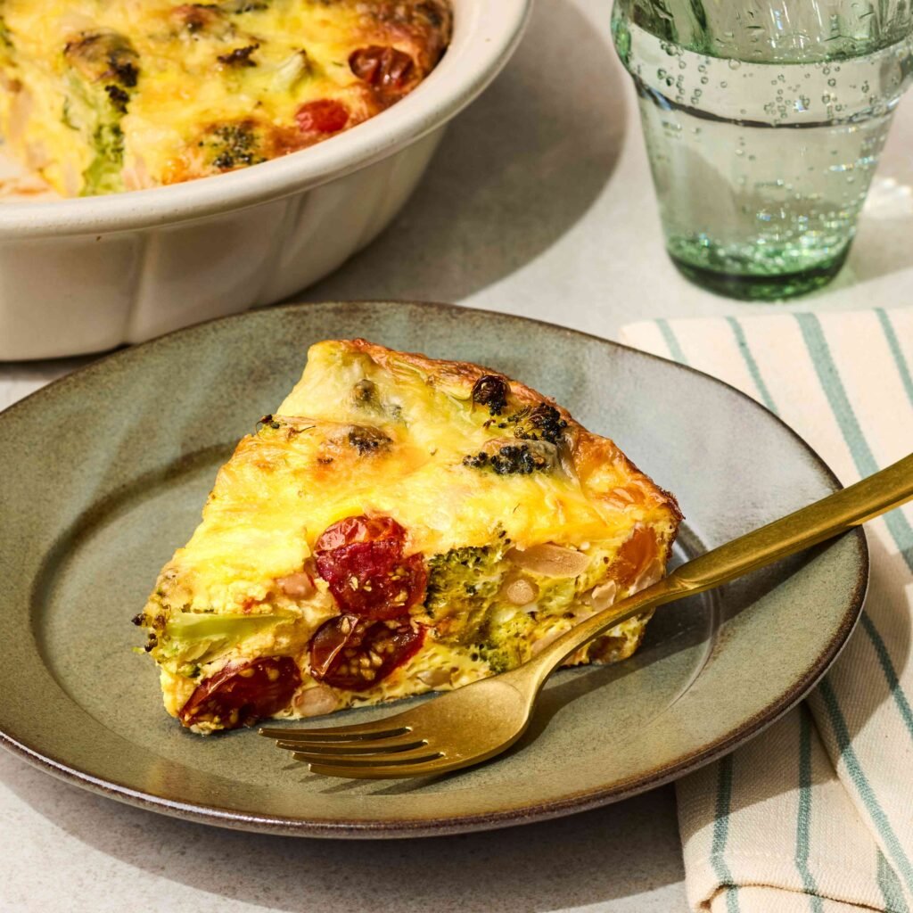 Fatia de quiche de legumes em prato cinza com garfo dourado ao lado de copo e pano listrado.