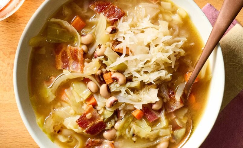 Tigela de sopa com vegetais, feijão, bacon e repolho sobre uma mesa de madeira.