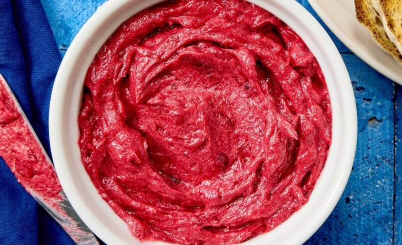 Creme de beterraba em tigela branca sobre uma superfície azul, com faca e torradas ao lado.