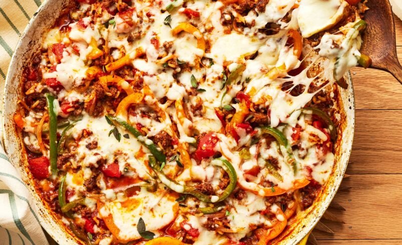 Frigideira amarela com carne moída, vegetais e queijo derretido preparada sobre mesa de madeira.