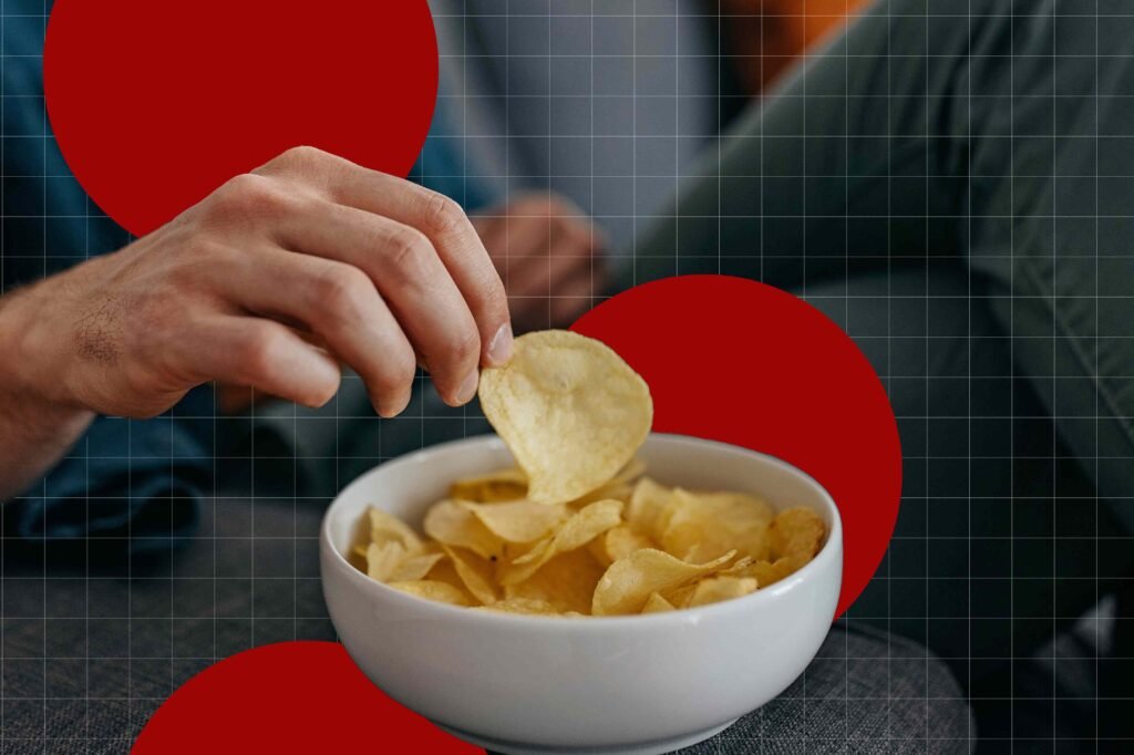 Close de mão pegando batata chip em tigela branca sobre superfície cinza.