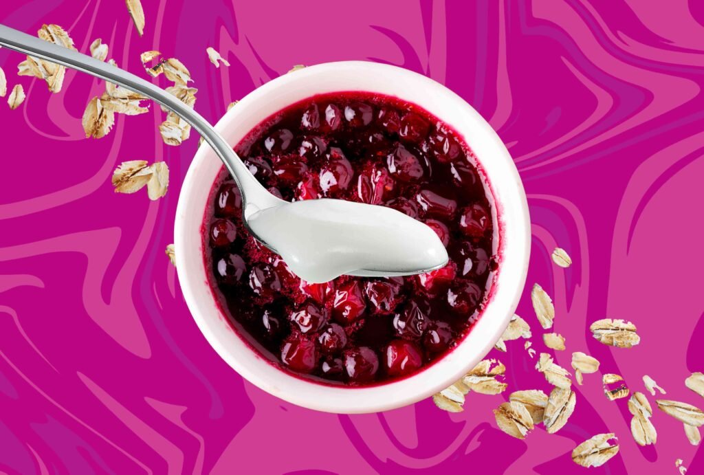 Tigela de iogurte com calda de frutas vermelhas e aveia sobre fundo colorido.
