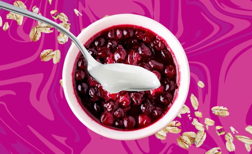 Tigela de iogurte com calda de frutas vermelhas e aveia sobre fundo colorido.
