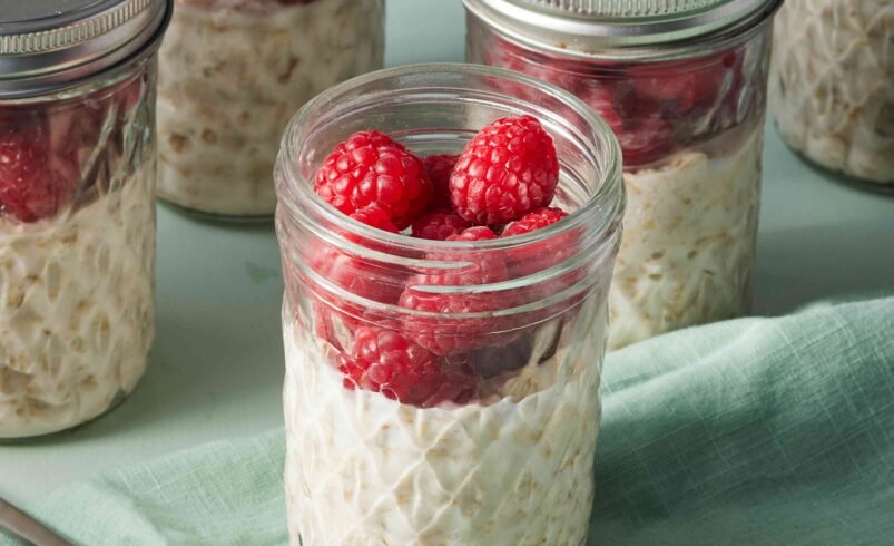 Potes de vidro contendo aveia e framboesas frescas sobre uma mesa verde.