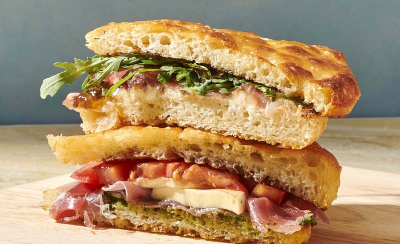 Dois pedaços de sanduíche com focaccia, presunto cru, rúcula, tomate e queijo sobre uma tábua de madeira.