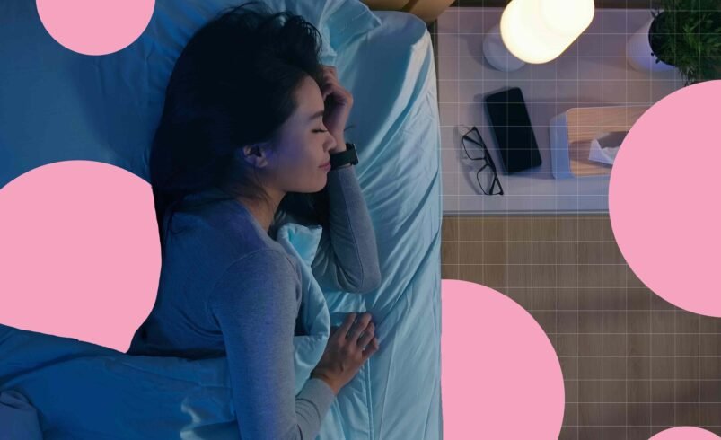 Pessoa deitada em cama com roupa cinza, luminária e mesa-de-cabeceira com itens.