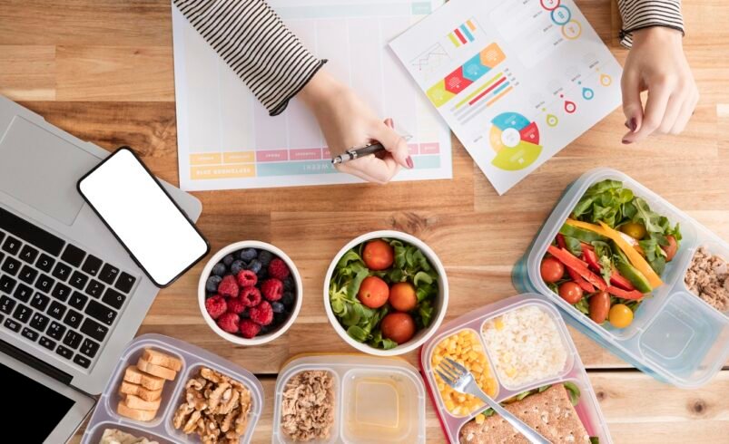 Mesa com laptop, celular, gráficos e refeições saudáveis organizadas em potes.