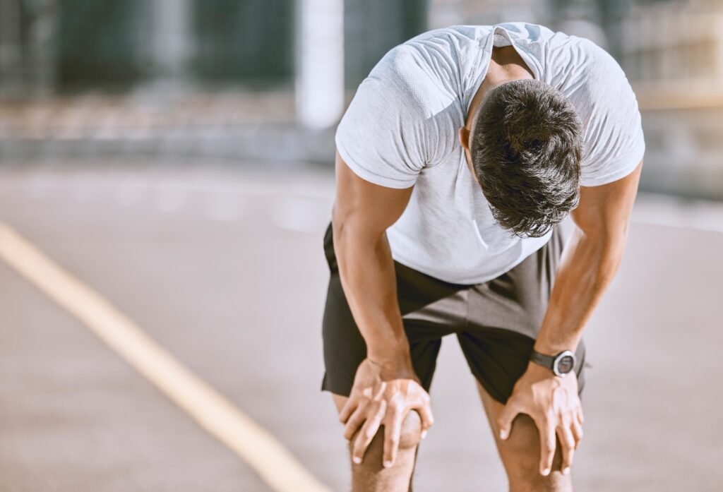 Homem de camiseta branca e shorts escuros inclinado, descansando após corrida.