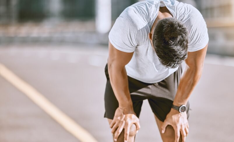 Homem de camiseta branca e shorts escuros inclinado, descansando após corrida.