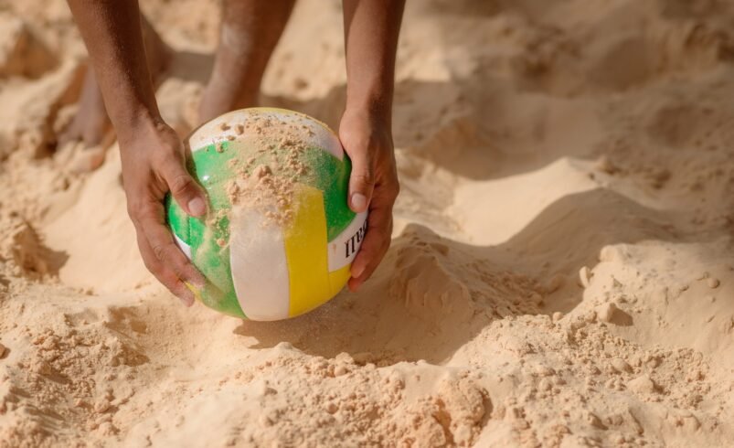 Mãos segurando uma bola de vôlei em uma quadra de areia.
