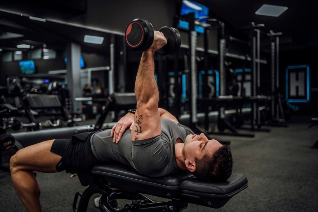 Homem deitado em banco de academia levantando haltere com uma mão.
