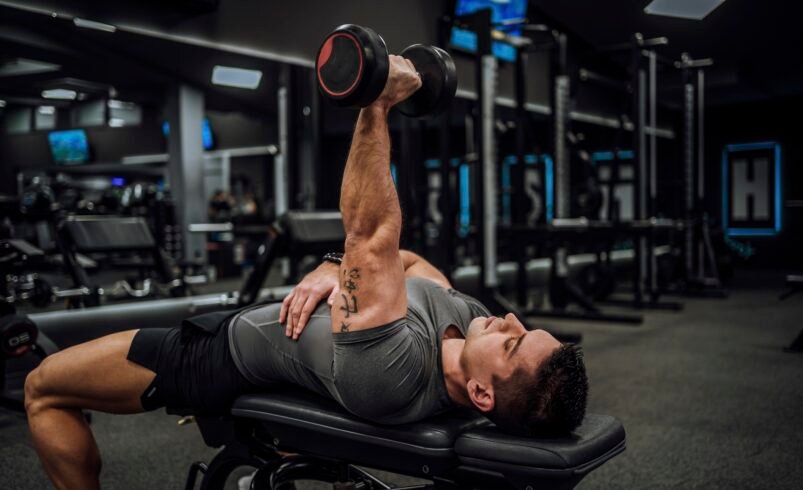 Homem deitado em banco de academia levantando haltere com uma mão.