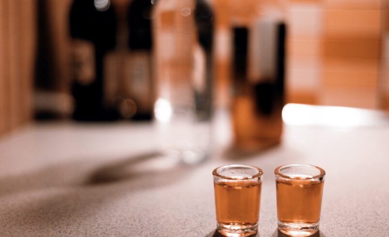 Dois copos de shot com bebida âmbar sobre balcão, garrafas desfocadas ao fundo.