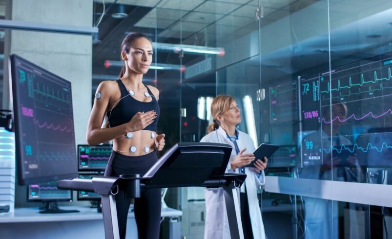 Pessoa correndo em esteira com sensores no corpo, acompanhada por profissional de saúde.