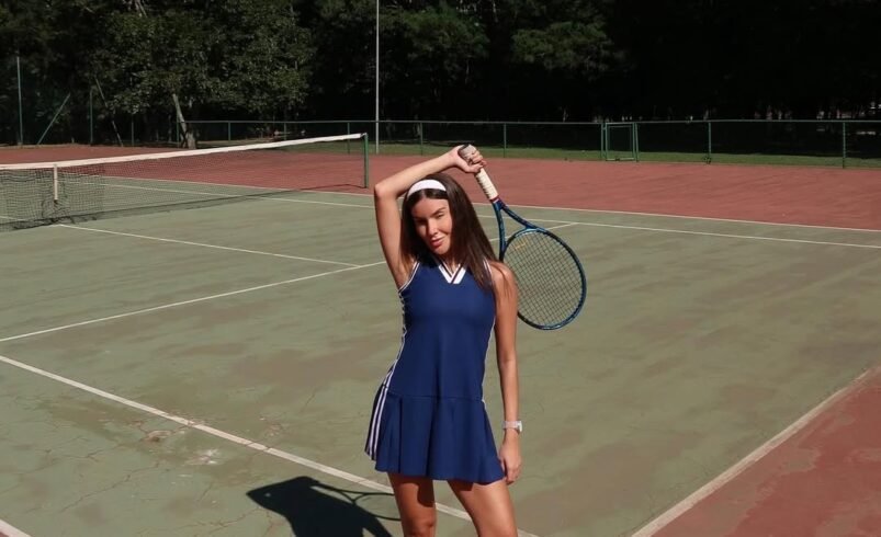 Pessoa em pé em quadra de tênis segura uma raquete sob o sol.