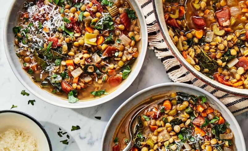 Sopa de lentilha servida em tigelas com legumes e queijo ralado ao lado.