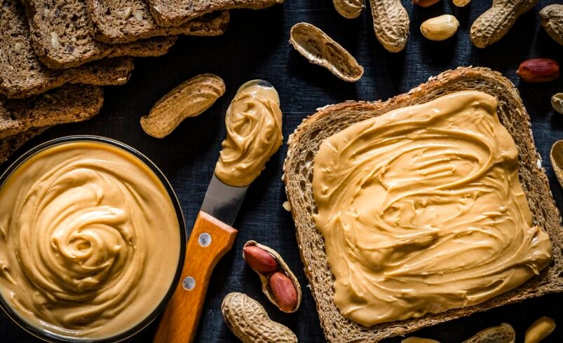 Pasta de amendoim espalhada em pão integral, amendoins inteiros e tigela com pasta.