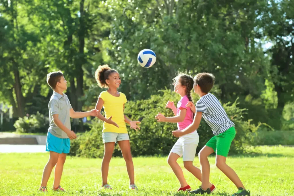 Quatro crianças brincando com bola em área verde em dia de sol.