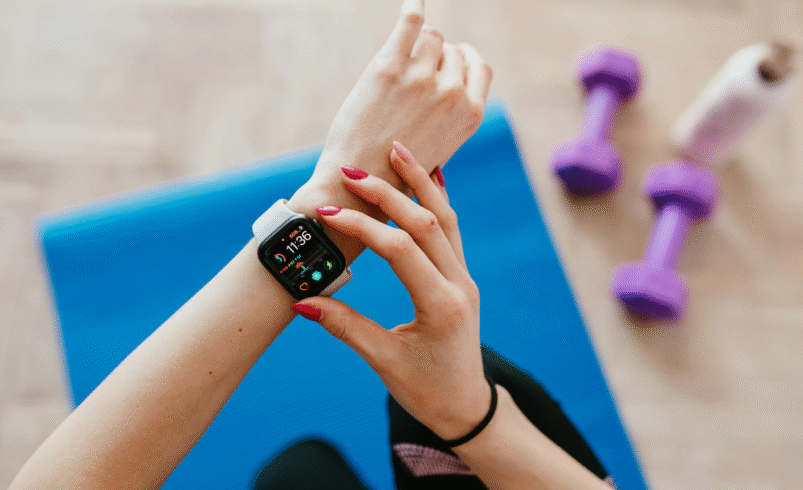 Mulher em tapete azul ajustando um smartwatch com pesos ao fundo.