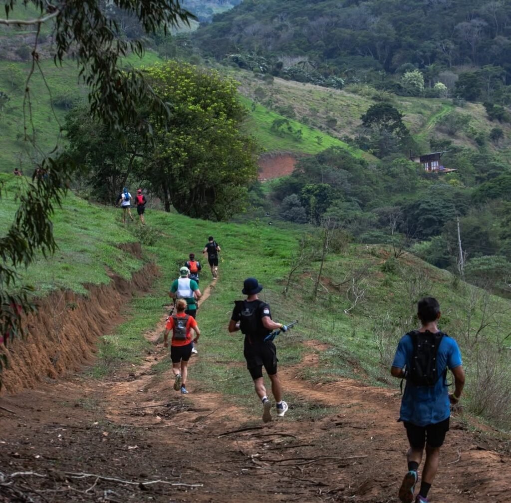 Grupo de corredores praticando trail running em trilha verde cercada por vegetação e colinas.
