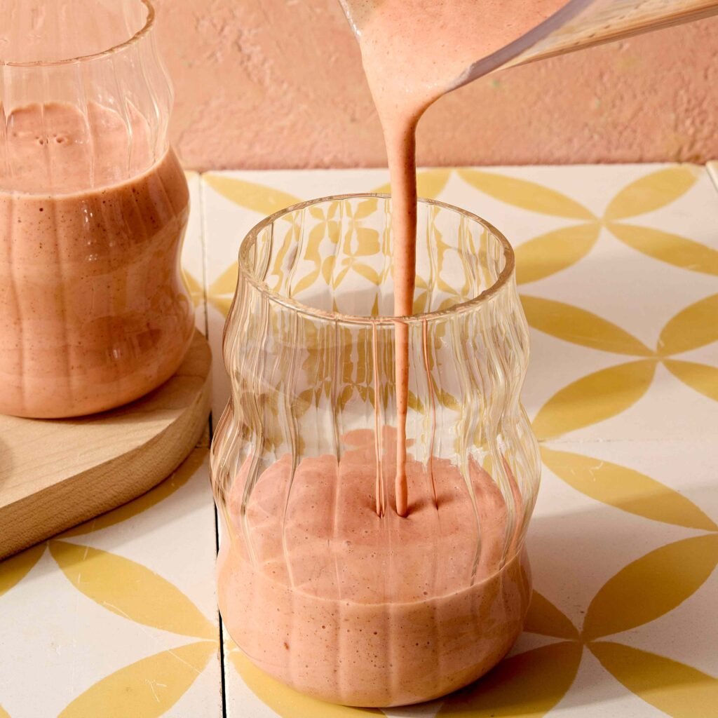 Smoothie cor-de-rosa sendo vertido em copo com design ondulado sobre uma superfície decorada.