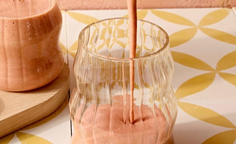 Smoothie cor-de-rosa sendo vertido em copo com design ondulado sobre uma superfície decorada.