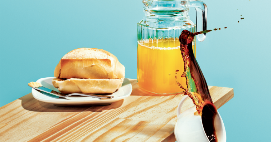 Pão francês com faca ao lado, suco de laranja em jarra e café sendo servido.