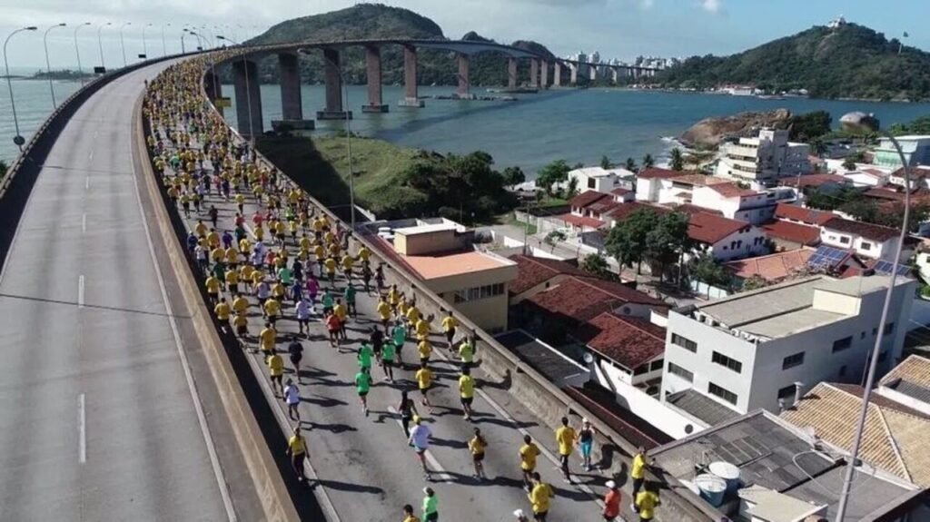 Corredores participam de maratona em ponte com vista para o mar e cidade ao fundo.