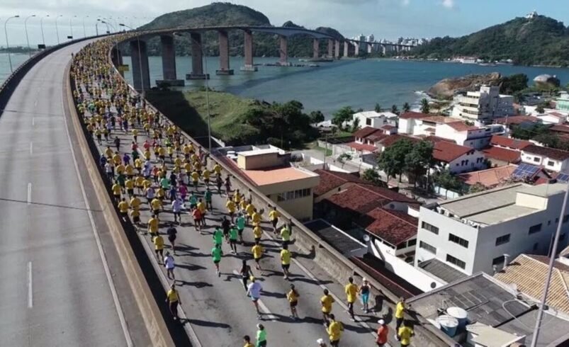 Corredores participam de maratona em ponte com vista para o mar e cidade ao fundo.