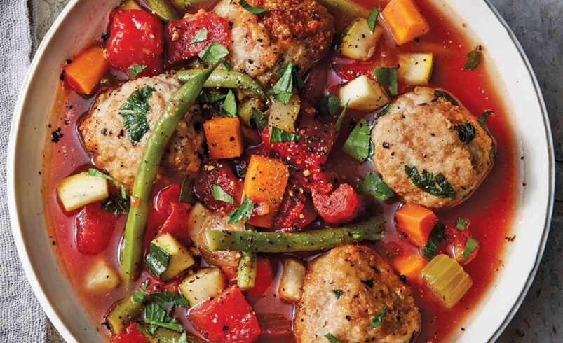 Tigela de sopa com almôndegas, legumes variados e caldo vermelho.