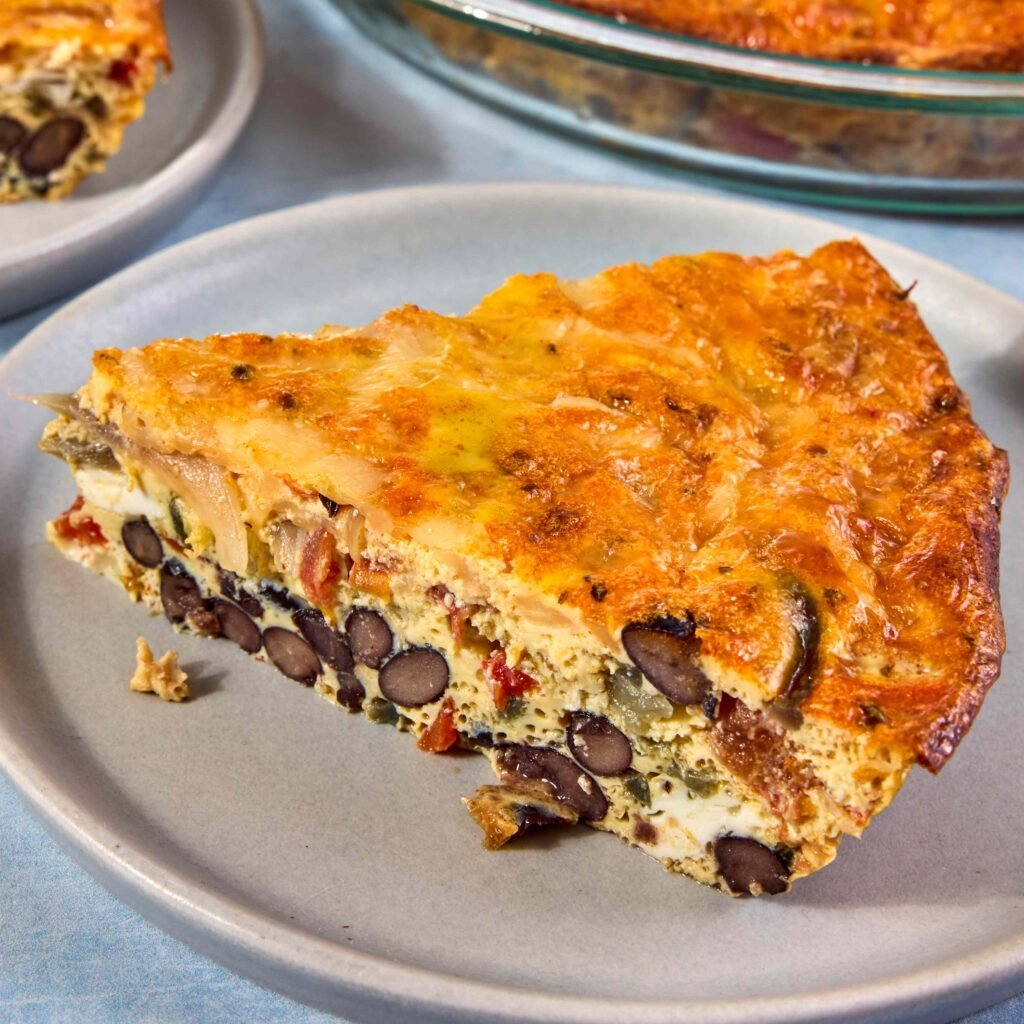 Fatia de quiche dourada com feijão preto e legumes em um prato cinza.