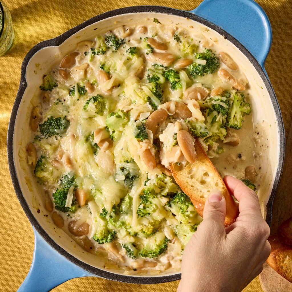 Mão mergulhando torrada em fondue de queijo com brócolis em panela azul.
