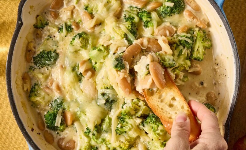 Mão mergulhando torrada em fondue de queijo com brócolis em panela azul.