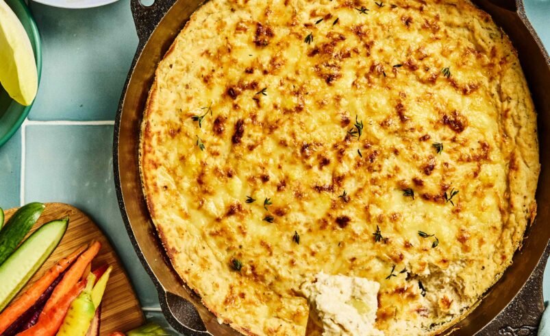 Travessa de queijo gratinado com legumes frescos e biscoitos ao redor.