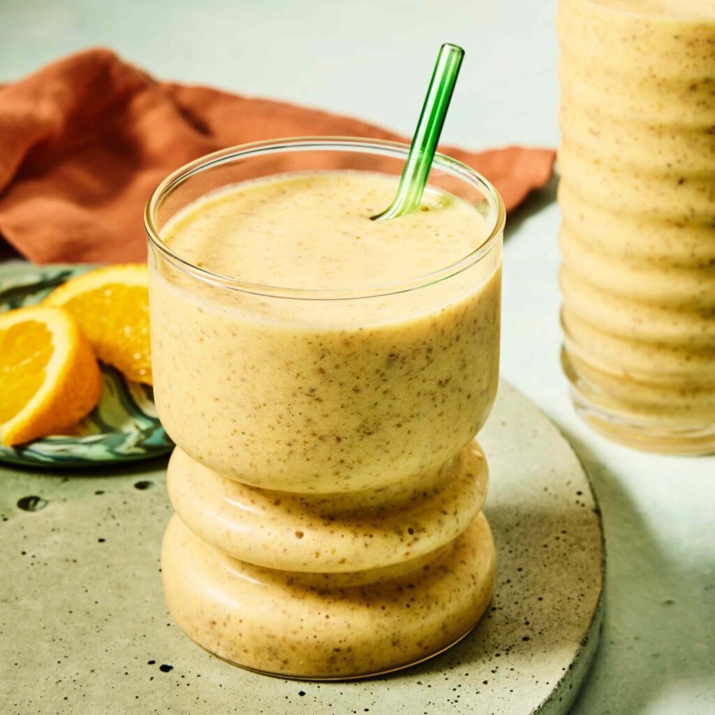 Smoothie cremoso de laranja em copo com canudo verde, prato com fatias de laranja ao fundo.