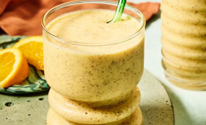 Smoothie cremoso de laranja em copo com canudo verde, prato com fatias de laranja ao fundo.