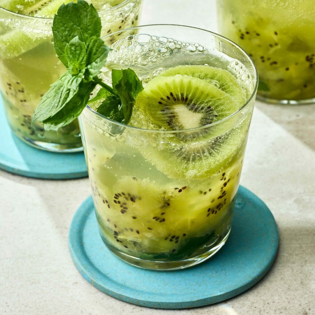 Mais de 10 bebidas saborosas para uma melhor saúde intestinal 1 Copo transparente com drink de kiwi decorado com hortelã em base azul.