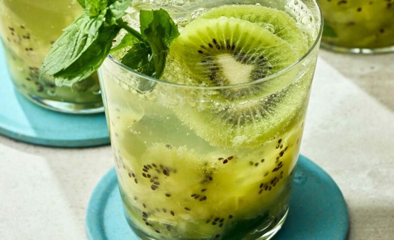 Copo transparente com drink de kiwi decorado com hortelã em base azul.