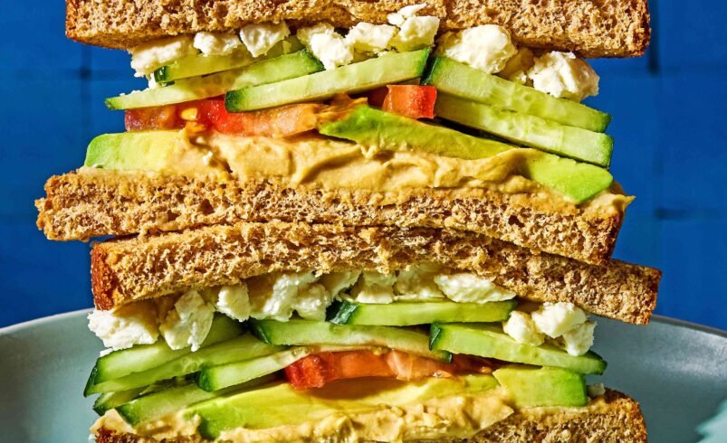 Sanduíche de pão integral com vegetais, queijo e patê em prato azul.