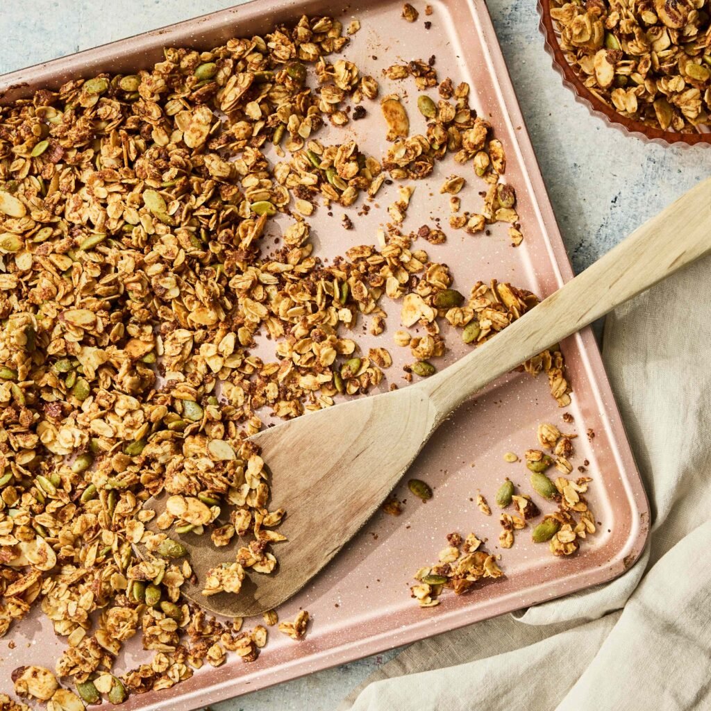 Granola dourada espalhada em uma assadeira rosa com colher de madeira.