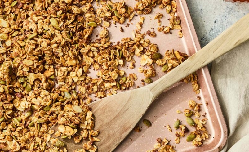 Granola dourada espalhada em uma assadeira rosa com colher de madeira.