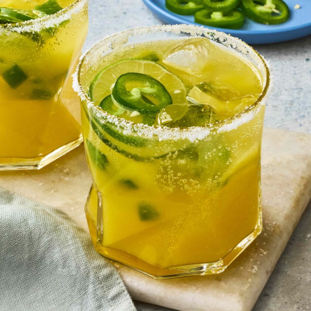 Um copo com drink amarelo, decorado com fatias de jalapeños e borda de sal.