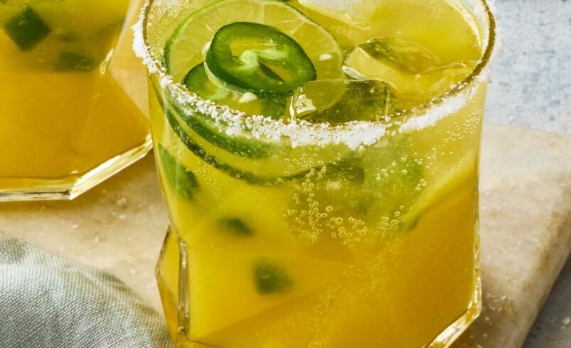 Um copo com drink amarelo, decorado com fatias de jalapeños e borda de sal.