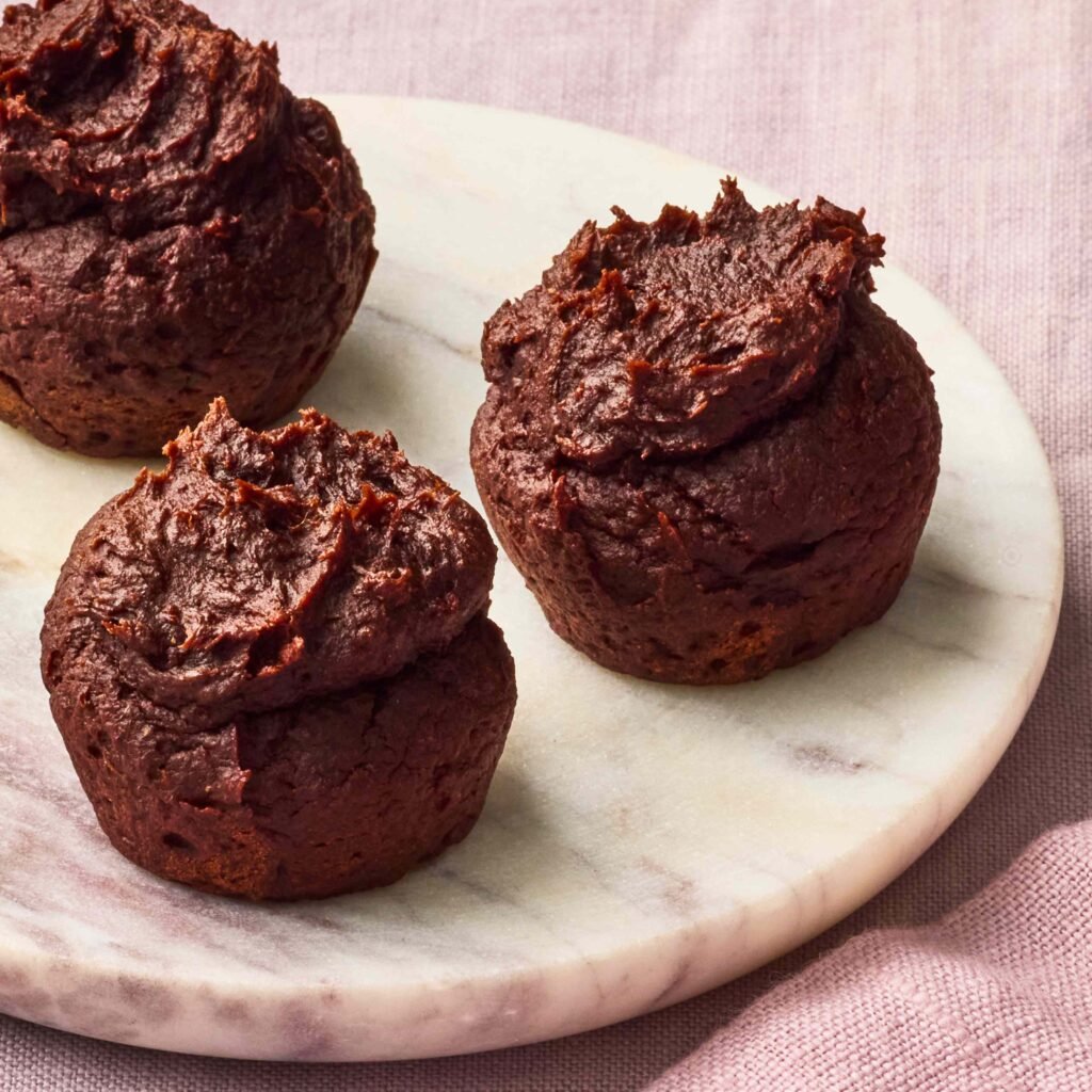 Três muffins de chocolate em prato de mármore com fundo lilás.