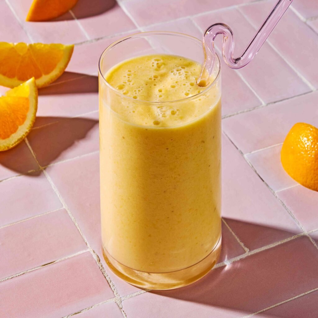 Copo de smoothie amarelo com canudo de vidro sobre mesa rosa e laranjas cortadas.