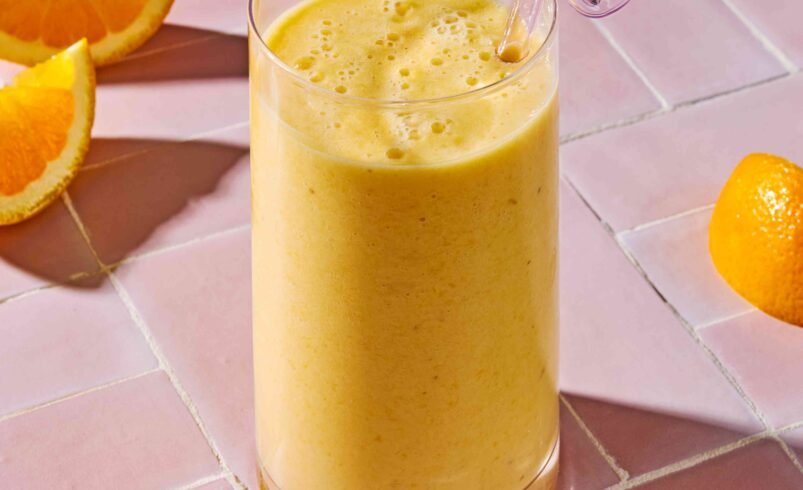 Copo de smoothie amarelo com canudo de vidro sobre mesa rosa e laranjas cortadas.